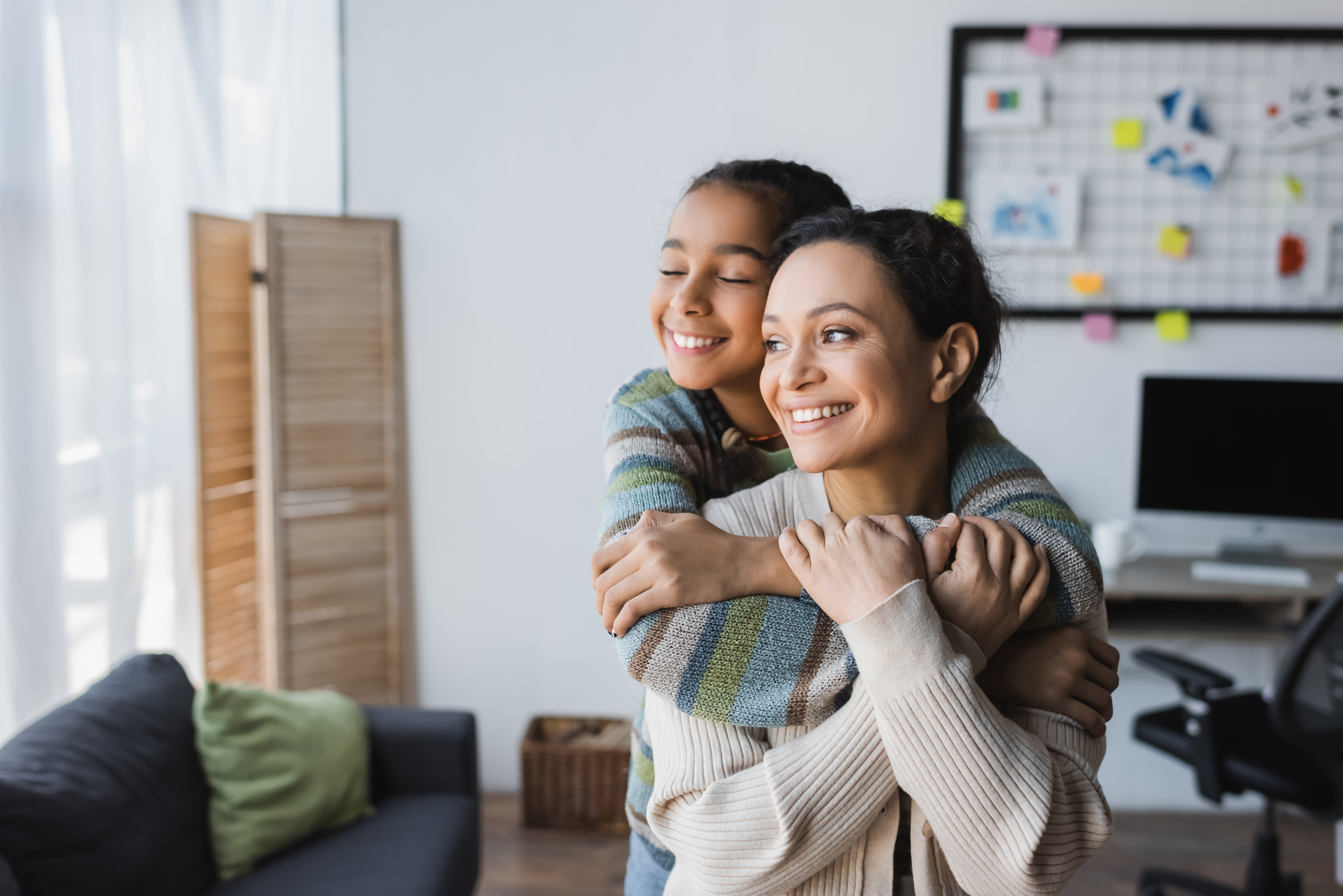 A young girl hugs her mom, who is smiling while sitting at a computer, illustrating a positive parent-child relationship with technology.